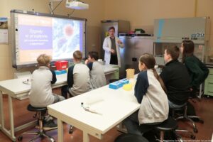 Noc Biologów 2026. Sala laboratoryjna. Ujęcie bokiem. Młodzież (tyłem do kadru) siedząca przy białych długich stołach ustawionych jeden za drugim. Leżą na nich przyrządy laboratoryjne. Po prawej w tle przodem do kadru laborant w białym fartuchu opowiadający o odporności. Po lewej ekran z prezentacją.