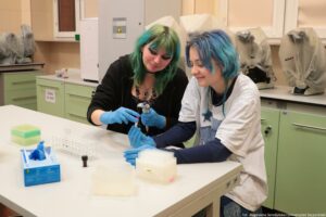 Noc Biologów 2026. Sala laboratoryjna. Dwie dziewczyny – jedna w niebieskich włosach, a druga w zielony – mają założone niebieskie lateksowe i siedzą przy długim białym stole, na którym leżą przyrządy laboratoryjne. Jedna z dziewczyn trzyma probówkę, a druga pipetę automatyczną.
