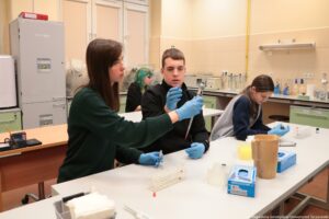 Noc Biologów 2026. Sala laboratoryjna. Ujęcie bokiem. Młodzież w niebieskich lateksowych rękawiczkach siedząca przy białych długich stołach ustawionych jeden za drugim. Leżą na nich przyrządy laboratoryjne. Na pierwszym planie dziewczyna trzymająca probówkę i przekazująca pipetę automatyczną swojemu koledze.