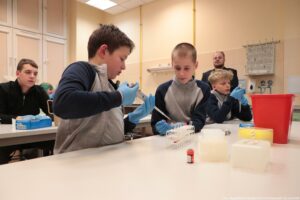 Noc Biologów 2026. Sala laboratoryjna. Ujęcie bokiem. Młodzież w niebieskich lateksowych rękawiczkach siedząca przy białych długich stołach ustawionych jeden za drugim. Leżą na nich przyrządy laboratoryjne. Na pierwszym planie chłopak, który pipety automatycznej wlewa preparat do probówki.