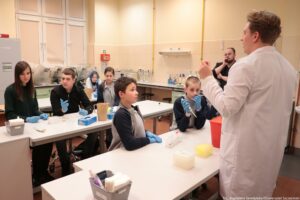 Noc Biologów 2026. Sala laboratoryjna. Młodzież w niebieskich lateksowych rękawiczkach siedząca przy białych długich stołach ustawionych jeden za drugim. Leżą na nich przyrządy laboratoryjne. Po prawej bokiem do kadru laborant w białym fartuchu tłumaczący przebieg eksperymentu.