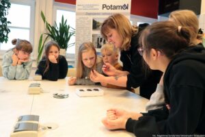Noc Biologów 2026. Duży biały stół, dookoła którego siedzą dzieci zaciekawione tym, co pokazuje im prowadząca. W tle biały roll up na temat zagrożenia dla wód