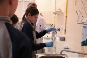 Noc Biologów 2026. Sala laboratoryjna. Ujęcie bokiem. Po prawej biały stół z przyrządami laboratoryjnymi, po lewej troje dzieci – ujęcie na dziewczynkę w niebieskich lateksowych rękawiczkach, która trzyma jeden z przyrządów.