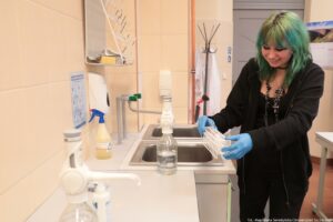 Noc Biologów 2026. Po prawej uśmiechnięta dziewczyna w zielonych włosach niesie biały statyw na probówki. Obok niej metalowy zlew dwukomorowy. W tle stojący wieszak z białymi fartuchami.