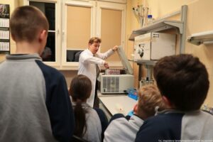 Noc Biologów 2026. Czworo dzieci (tyłem do kadru). W tle laborant w białym fartuchu otwierający specjalistyczne urządzenie. Za nim okna z beżowymi roletami.