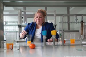 Noc Biologów 2026. Sala laboratoryjna. Na wprost siedząca przy stole laborantka wlewa coś do kubka z sokiem z mandarynek.