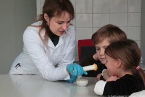 Noc Biologów 2026. Sala laboratoryjna. Po prawej dwoje dzieci, po lewej laborantka oparta o stół pokazuje dzieciom, co odgniotła w moździerzu.