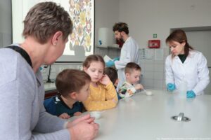 Noc Biologów 2026. Sala laboratoryjna. Czworo dzieci i ich opiekunka (po lewej) przygotowują się do eksperymentu. Pomagają im osoby z obsługi (kobieta przy stole i mężczyzna za nimi).