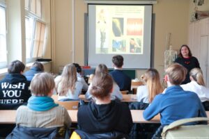 Noc Biologów 2026. Sala wykładowa. Uczniowie w ławkach tyłem do kadru. Prelegentka po prawej stronie prezentuje wykład. Pośrodku na ekranie slajd związany z anatomią.