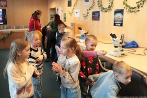 Noc Biologów 2026. Korytarz. Siedmioro dzieci rozmawia obok stołu z mikroskopami.