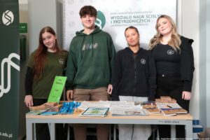 Noc Biologów 2026. Przedstawiciele Rady Samorządu Studenckiego Wydziału Nauk Ścisłych i Przyrodniczych w koszulkach i bluzach z logiem Uniwersytetu Szczecińskiego przy stoisku z elementami gry terenowej organizowanej w ramach Nocy Biologów 2026.