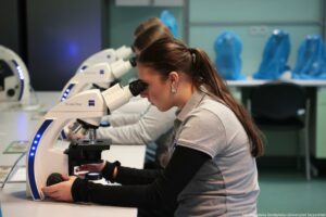 Noc Biologów 2026. Sala laboratoryjna i osoby obserwujące preparaty pod mikroskopami (ujęcie bokiem).