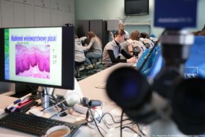 Noc Biologów 2026. Na pierwszym planie po lewej stronie monitor z napisem – Nabłonek wielowarstwowy płaski (język kota) i zdjęcie w powiększeniu. W tle osoby obserwujące preparaty pod mikroskopami.