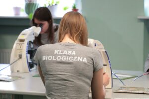 Noc Biologów 2026. Na pierwszym planie tyłem do kadru młoda kobieta patrząca w mikroskop. Na szarej koszulce ma biały napis – klasa biologiczna. W tle (przodem do kadru) druga kobieta patrząca w mikroskop.