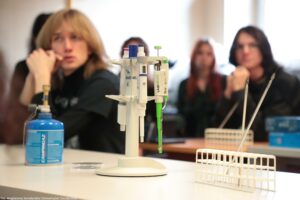 Noc Biologów 2026. Na środku stołu laboratoryjne pipety na stojaku. W tle uczniowie.