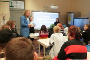 Noc Biologów 2026. Laboratoryjna sala wykładowa. Tyłem do kadru uczniowie siedzący za stolikami. W tle prowadzący i napis na ekranie: Jakie mikroorganizmy są wokół nas?