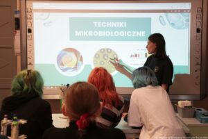 Noc Biologów 2026. Na pierwszym planie tyłem do kadru cztery uczennice. W tle wykładowczyni przed ekranem z napisem: „Techniki mikrobiologiczne”.