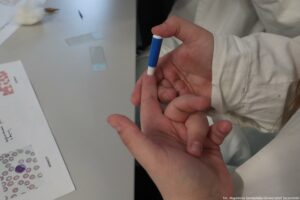 Noc Biologów 2026. Prawa dłoń nakłuwa lewy palec wskazujący. W tle na stole szkiełka mikroskopowe.