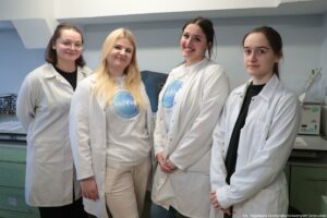 Noc Biologów 2026. Cztery laborantki w białych fartuchach uśmiechają się do zdjęcia, stojąc w laboratorium na tle sprzętów laboratoryjnych.
