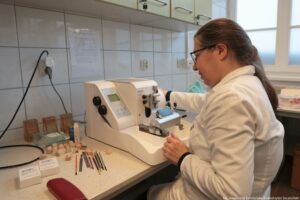 Noc Biologów 2026. Laboratorium. Kobieta siedząca tyłem w białym fartuchu pracuje przy stojącym na stole sprzęcie – mikrotomie rotacyjnym. Obok niego po lewej stronie leżą m.in. pędzelki i nożyki.