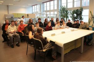 Noc Biologów 2026. Laboratorium. Ponad 20-osobowa grupa z uczniami siedzi przy dużym stole i słucha prelekcji. W tle szafki i półki z przyrządami laboratoryjnymi.