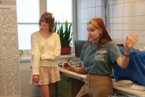 Noc Biologów 2026. Dwie młode kobiety w laboratorium. Kobieta w tle stoi przodem do kadru i słucha prelekcji. Kobieta z przodu (bokiem do kadru) omawia temat jezior.