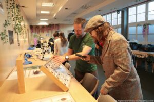Noc Biologów 2026. Korytarz. Na pierwszym planie po lewej stronie pod ścianą stoi bokiem ławka, na niej gabloty z owadami, motylami, w tle mikroskopy. Przy ławce stoi bokiem dwóch mężczyzn i oglądają jedną z gablot.