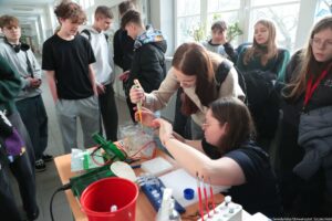 Noc Biologów 2026. Korytarz. Na pierwszym planie bokiem ławka z przyrządami. Za nią dwie młode kobiety uzupełniające probówki. Dookoła uczniowie obserwujący proces.
