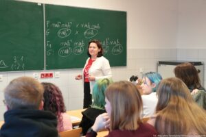 Noc Biologów 2026. Sala lekcyjna. Na pierwszym planie uczniowie w ławkach (tyłem do kadru). W tle nauczycielka przed zieloną tablicą.