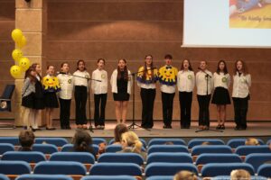 Grupa nastolatków stojących w szeregu na scenie i śpiewających do mikrofonów. Mają białe koszule z okrągłymi emblematami na piersi oraz ciemne spodnie lub spódnice; część ma na sobie żółto-niebieskie pelerynki z dużą uśmiechniętą buźką. Po lewej stronie sceny stoją żółte balony z narysowanymi uśmiechami. W tle jest brązowa ściana sali, a w prawym górnym rogu widać fragment wyświetlanego slajdu z częściowym napisem. Na pierwszym planie znajdują się rzędy niebieskich foteli i kilka osób siedzących na widowni. W prawym dolnym rogu widać mały podpis fotograficzny: „fot. Magdalena Seredyńska/Uniwersytet Szczeciński”.