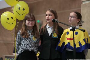 Trzy dziewczynki stojące obok mikrofonu, a po lewej stronie trzy żółte balony z czarnymi uśmiechniętymi buźkami. Potem widać, że scena jest w pomieszczeniu, na tle beżowej ściany. Dziewczynka po lewej ma jasną skórę, długie jasnobrązowe włosy z grzywką i okrągłe okulary. Ma biało-czarną bluzkę we wzór i czarne spodnie. Na piersi ma przypiętą okrągłą plakietkę z żółto-niebieskim motywem. Dziewczynka w środku ma jasną skórę i długie jasnobrązowe włosy, rozpuszczone. Ma białą koszulę i czarną marynarkę, a na piersi podobną okrągłą plakietkę. Dziewczynka po prawej ma jasną skórę, ciemne włosy związane do tyłu i okrągłe okulary. Ma na sobie żółty „pelerynowy” element stroju z dużą uśmiechniętą buźką i niebieskimi kształtami, pod spodem białą koszulę. Przed nią przebiega ramię statywu mikrofonowego. W prawym dolnym rogu widoczny napis: „fot. Magdalena Seredyńska/Uniwersytet Szczeciński”.