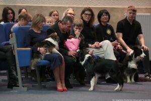 Grupa osób siedzących w rzędzie na niebieskich fotelach w sali audytoryjnej, z kilkoma psami rasy border collie na pierwszym planie. W pierwszym rzędzie siedzi kilka osób w ciemnych ubraniach; jedna kobieta w granatowej sukience ma na kolanach psa, obok mężczyzna trzyma różowy bukiet kwiatów, a obok nich siedzi chłopiec z ręką przy twarzy. Na podłodze przed siedzącymi stoi czarno-biały border collie zwrócony w ich stronę, a po prawej stronie kolejny czarno-biały pies siedzi przy młodym mężczyźnie w czarnej koszulce. W tle widać więcej osób, w tym kilka kobiet, a także fragment kaloryfera przy prawej krawędzi. W prawym dolnym rogu jest mały podpis: „fot. Magdalena Seredyńska/Uniwersytet Szczeciński”.