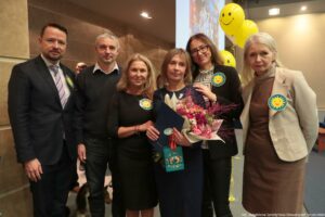 Uroczystość wręczenia Orderu Uśmiechu. Laureatka, trzymająca kwiaty, pozuje do zdjęcia z pracownikami uczelni. W tle żółte balony z uśmiechami.