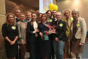 Uroczystość wręczenia Orderu Uśmiechu. Laureatka, trzymająca kwiaty, pozuje do zdjęcia z pracownikami uczelni. W tle żółte balony z uśmiechami.