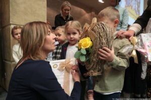 Uroczystość wręczenia Orderu Uśmiechu. Ujęcie bokiem. Laureatka kuca otoczona dziećmi, które wręczają jej dyplom i wiklinowy wieniec z żółtą różą.
