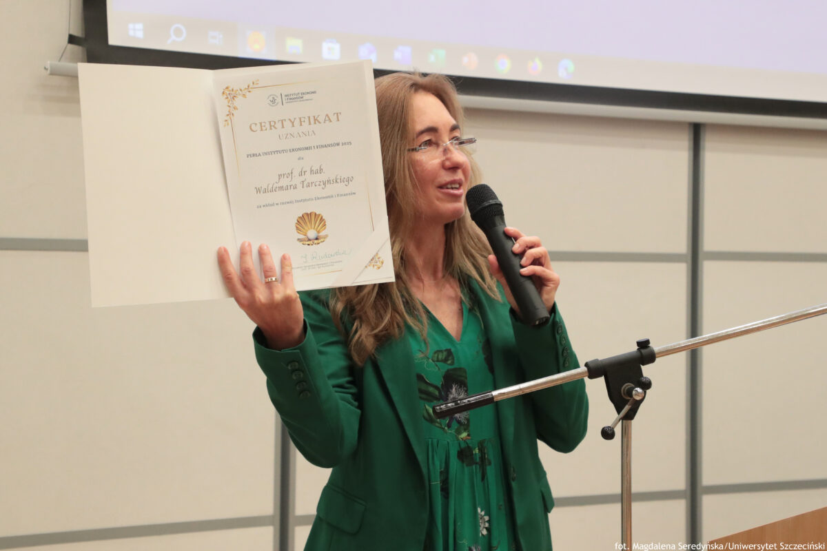 Za mównicą dyrektorka Instytutu Ekonomii i Finansów otwiera uroczystość wręczenia Pereł Instytutu Ekonomii i Finansów 2025 i prezentuje certyfikat w teczce.