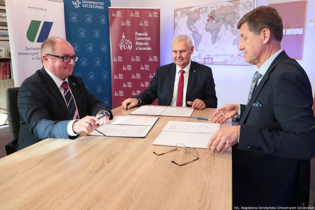 Trzech mężczyzn w garniturach przy drewnianym stole z dokumentami do podpisu. Dwóch siedzi po lewej i na środku, a trzeci stoi lub opiera się po prawej; na stole leżą otwarte teczki z kartkami oraz długopisy, a z przodu widać parę okularów. W tle stoją pionowe banery uczelni z nazwami: „Zachodniopomorski Uniwersytet Technologiczny w Szczecinie” (z adresem „www.zut.edu.pl”), „Uniwersytet Szczeciński” oraz „Pomorski Uniwersytet Medyczny w Szczecinie (PUM)”. Na ścianie po prawej wisi duża mapa świata. Podpis w prawym dolnym rogu: „fot. Magdalena Seredyńska/Uniwersytet Szczeciński”.