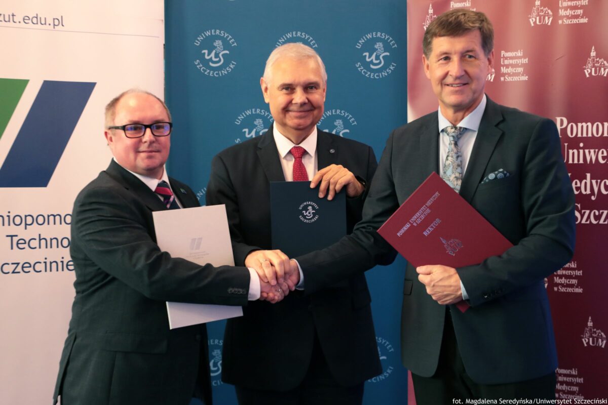 Trzech mężczyzn w garniturach stojących przed trzema ściankami z logotypami uczelni, z uściskiem dłoni na pierwszym planie. Wszyscy mają jasną karnację. Mężczyzna po lewej ma krótkie, przerzedzone włosy, okulary i ciemny garnitur z koszulą oraz krawatem w paski; trzyma białą teczkę/dokument. Mężczyzna w środku ma krótkie siwe włosy, ciemny garnitur i czerwony krawat; trzyma granatową teczkę z napisem i logo „UNIWERSYTET SZCZECIŃSKI”. Mężczyzna po prawej jest wyższy, ma brązowe włosy, ciemny garnitur, jasną koszulę i wzorzysty krawat; trzyma bordową teczkę z napisem „POMORSKI UNIWERSYTET MEDYCZNY W SZCZECINIE” oraz „REKTOR”. W tle, na niebieskiej ściance, powtarza się tekst „UNIWERSYTET SZCZECIŃSKI” z logo. Na bordowej ściance w tle powtarza się „Pomorski Uniwersytet Medyczny w Szczecinie” oraz skrót „PUM”. Na dole po prawej jest podpis: „fot. Magdalena Seredyńska/Uniwersytet Szczeciński”.