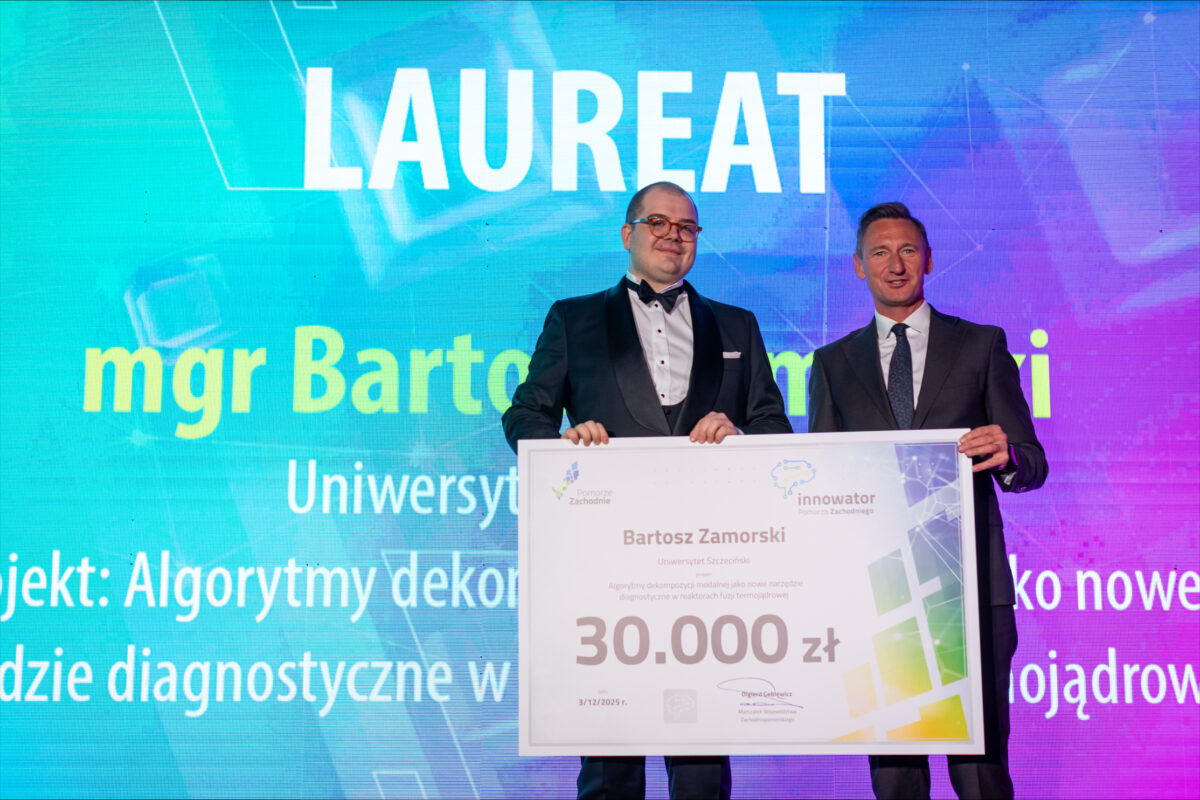 Dwóch mężczyzn w garniturach na scenie, trzymających duży, poziomy dyplom-czek, na tle kolorowego ekranu z dużym napisem „LAUREAT”. Mężczyzna po lewej ma czarny smoking z muchą i okulary; mężczyzna po prawej ma ciemny garnitur z krawatem. Na tle ekranu widać duże żółte litery zaczynające się od „mgr Barto…”, a niżej fragment „Uniwersy…”. Na dole tła widać urwany tekst zaczynający się od „Projekt: Algorytmy deko…” oraz kolejne niepełne linie tekstu.