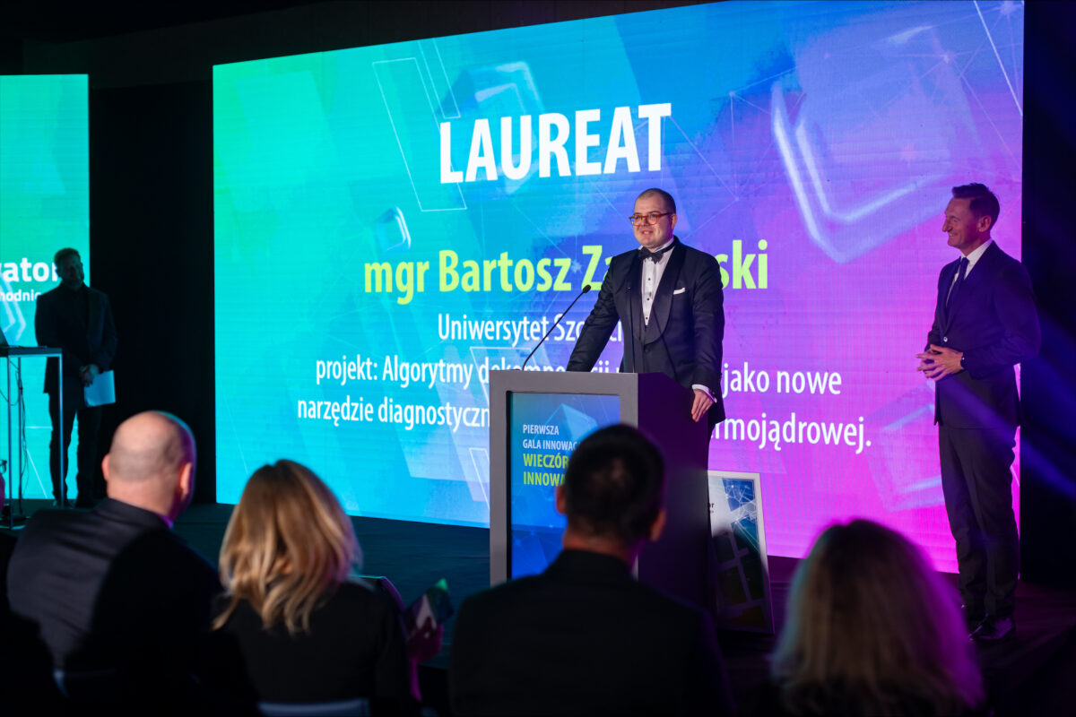 Scena z gali lub konferencji z dużym ekranem LED w tle i mównicą na środku. Na ekranie widoczny jest duży napis „LAUREAT”. Poniżej widać imię i nazwisko zaczynające się od „mgr Bartosz Z…”, ale dalsza część nazwiska jest nieczytelna na tym obrazie. Niżej znajduje się linia z „Uniwersytet …” (dalszy fragment jest nieczytelny) oraz tekst zaczynający się od „projekt: Algorytmy …” i dalsze zdania o narzędziu diagnostycznym; większość tych mniejszych napisów jest zbyt rozmazana, by przepisać je dokładnie. Na środku stoi mężczyzna w smokingu z czarną muchą, w okularach, przemawia przy mównicy. Po prawej stronie stoi drugi mężczyzna w ciemnym garniturze, uśmiechnięty, z rękami złożonymi z przodu, jak prowadzący lub wręczający nagrodę. Z przodu kadru widać tyły głów kilku osób siedzących na widowni. Na froncie mównicy jest panel z napisem zaczynającym się od „PIERWSZA GALA INNOWA…” oraz większymi słowami „WIECZÓR INNOWA…”, ale reszta również jest częściowo nieczytelna. Dominują niebiesko-turkusowo-fioletowe światła sceniczne.