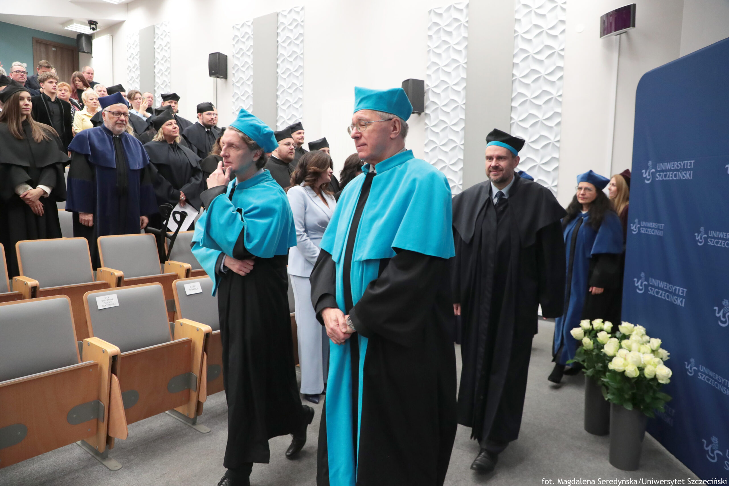 Uroczystość akademicka w auli z osobami w togach i czapkach akademickich. W centrum stoją dwie osoby w czarnych togach z turkusowymi pelerynami i turkusowymi biretami; jedna ma rękę przy twarzy, druga stoi z rękami złożonymi z przodu. Wokół nich znajdują się inni uczestnicy w czarnych togach, część z granatowymi elementami, stojący w rzędach przy ścianie. Po lewej stronie widać rzędy jasnoszarych, składanych foteli z drewnianymi podłokietnikami; na kilku siedzeniach są białe kartki z rezerwacją. Po prawej stronie stoi granatowa ścianka z powtarzającym się napisem „UNIWERSYTET SZCZECIŃSKI” i logo. Obok niej znajduje się wazon z białymi kwiatami. Na dole po prawej jest podpis: „fot. Magdalena Seredyńska/Uniwersytet Szczeciński”.