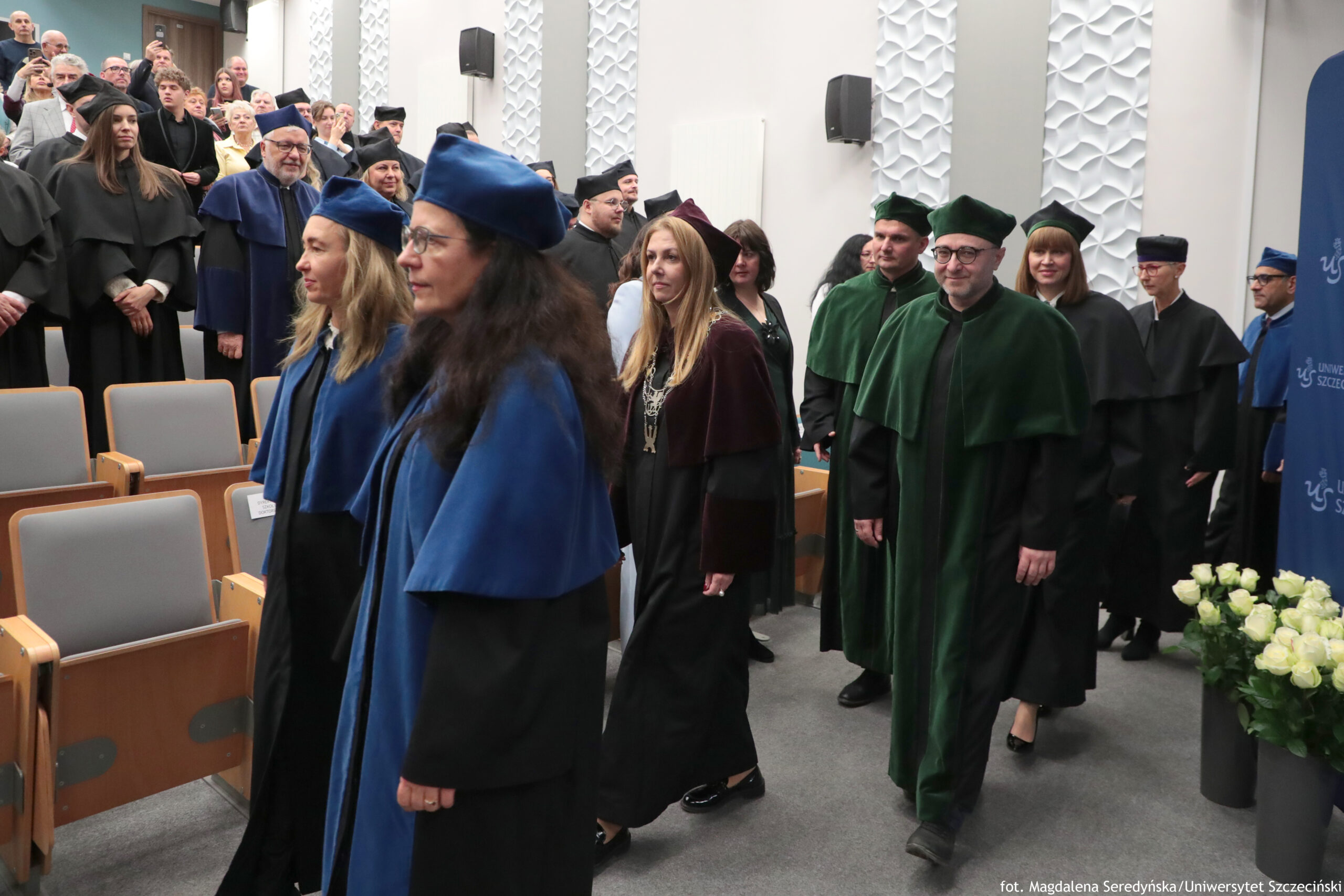 Grupa osób w strojach akademickich w auli, idąca w procesji między rzędami siedzeń. W pierwszym planie widać dwie kobiety w granatowych togach i granatowych biretach. Za nimi idą kolejne osoby w togach w różnych kolorach, m.in. zielonych, czarnych i bordowych; część ma birety, część czapki akademickie. Po lewej stronie, na podwyższeniu, stoi tłum osób w formalnych ubraniach i togach, obserwujących wydarzenie. Po prawej stronie jest niebieski pionowy baner z częściowo widocznym napisem „Uniwersytet Szczeciński” oraz wazon z białymi kwiatami. Na dole po prawej widoczny jest podpis fotograficzny: „fot. Magdalena Seredyńska/Uniwersytet Szczeciński”.
