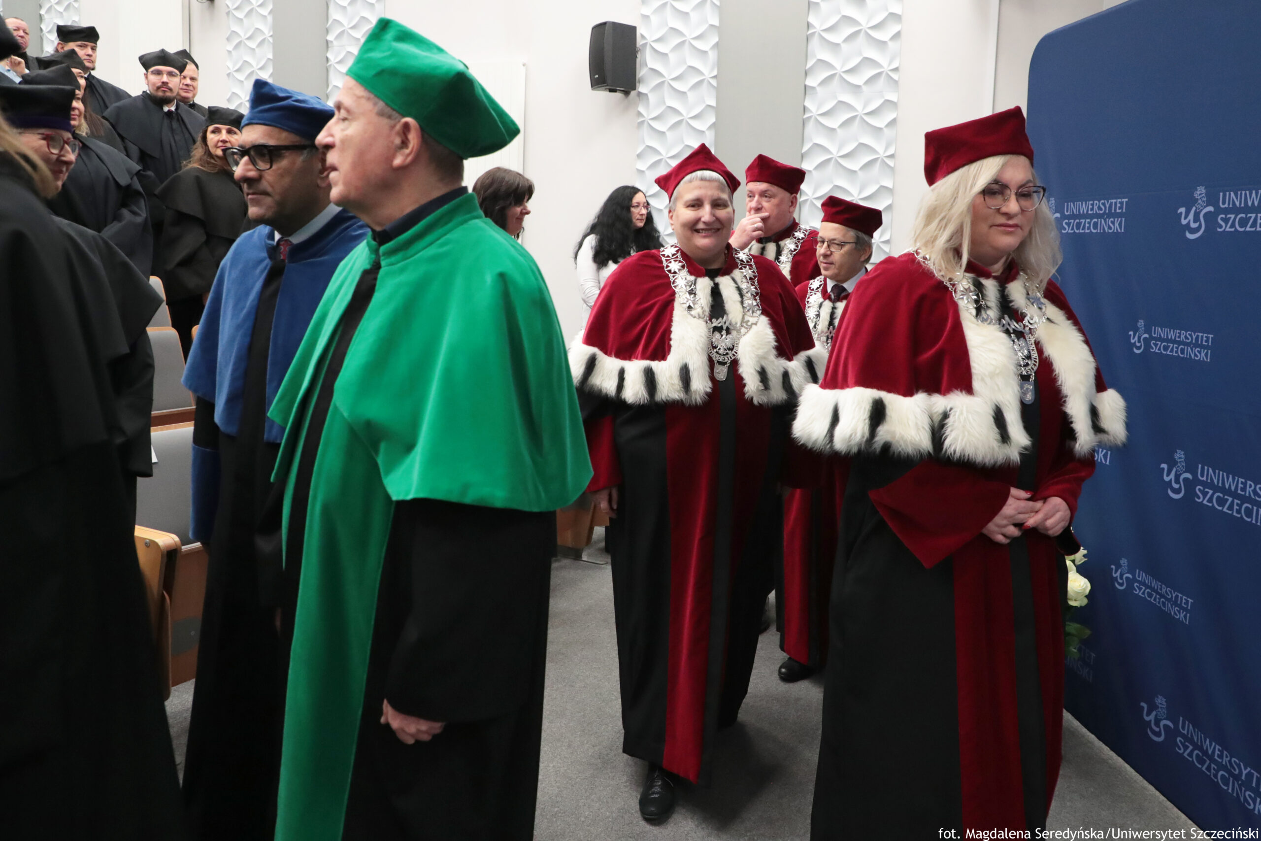 Grupa osób w strojach akademickich podczas uroczystości na uczelni. Widać kilka osób w togach i biretach: z przodu mężczyzna w zielonej todze i zielonym birecie, obok niego mężczyzna w niebieskiej todze i niebieskim birecie, a po prawej kilka osób w czerwonych togach z białym futrzanym obszyciem i czerwonych biretach. Z tyłu stoją kolejne osoby w czarnych togach. Po prawej stronie jest granatowa ścianka z powtarzającym się napisem „UNIWERSYTET SZCZECIŃSKI” oraz logo uczelni. W tle widać jasną ścianę z dekoracyjną, wypukłą fakturą i fragmenty sali z siedzeniami.