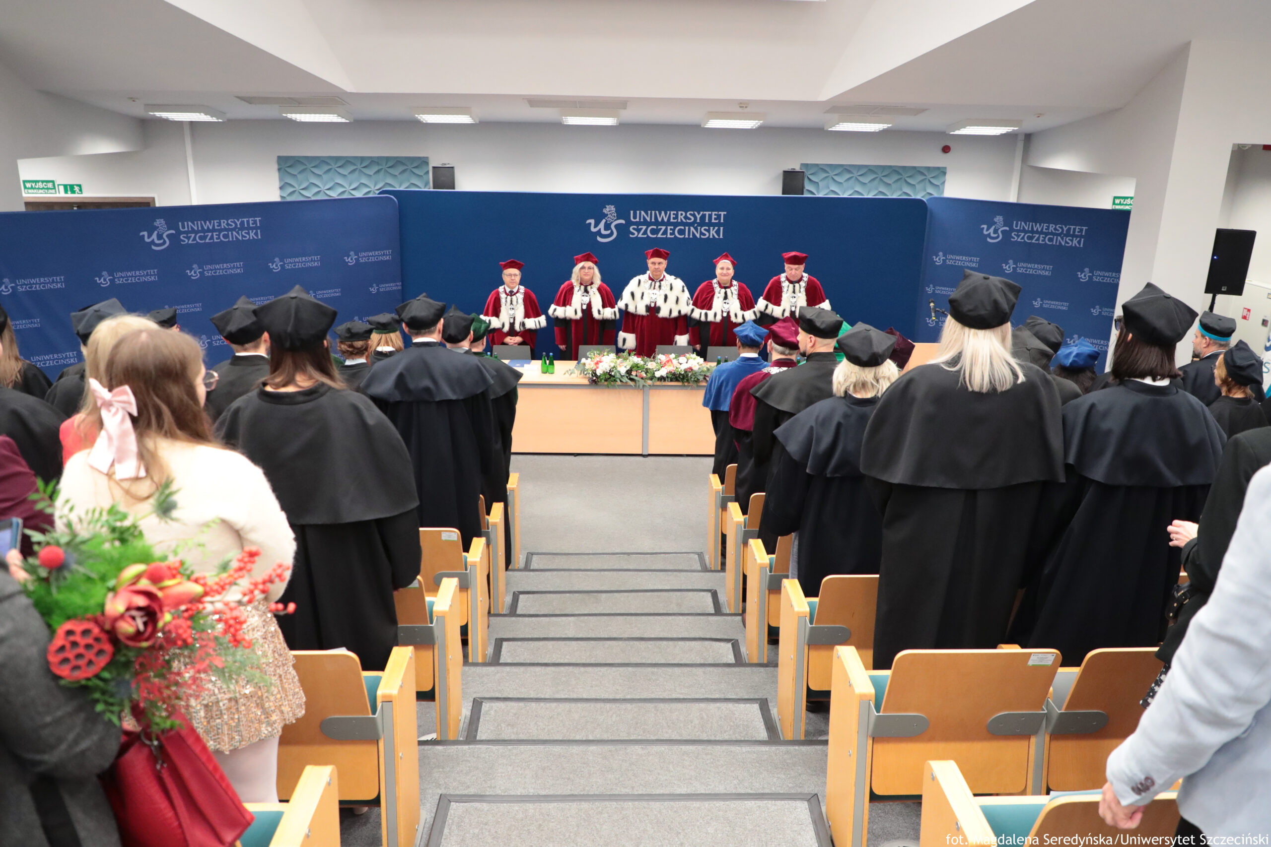 Sala wykładowa podczas uroczystości uniwersyteckiej. Na pierwszym planie widać rzędy drewnianych siedzeń i stojących uczestników w czarnych togach oraz biretach. Z przodu sali, za długim stołem z kompozycją kwiatową, stoi pięć osób w czerwono-białych, ceremonialnych strojach akademickich. Tło stanowią niebieskie ścianki z powtarzającym się napisem „UNIWERSYTET SZCZECIŃSKI” oraz logo uczelni.