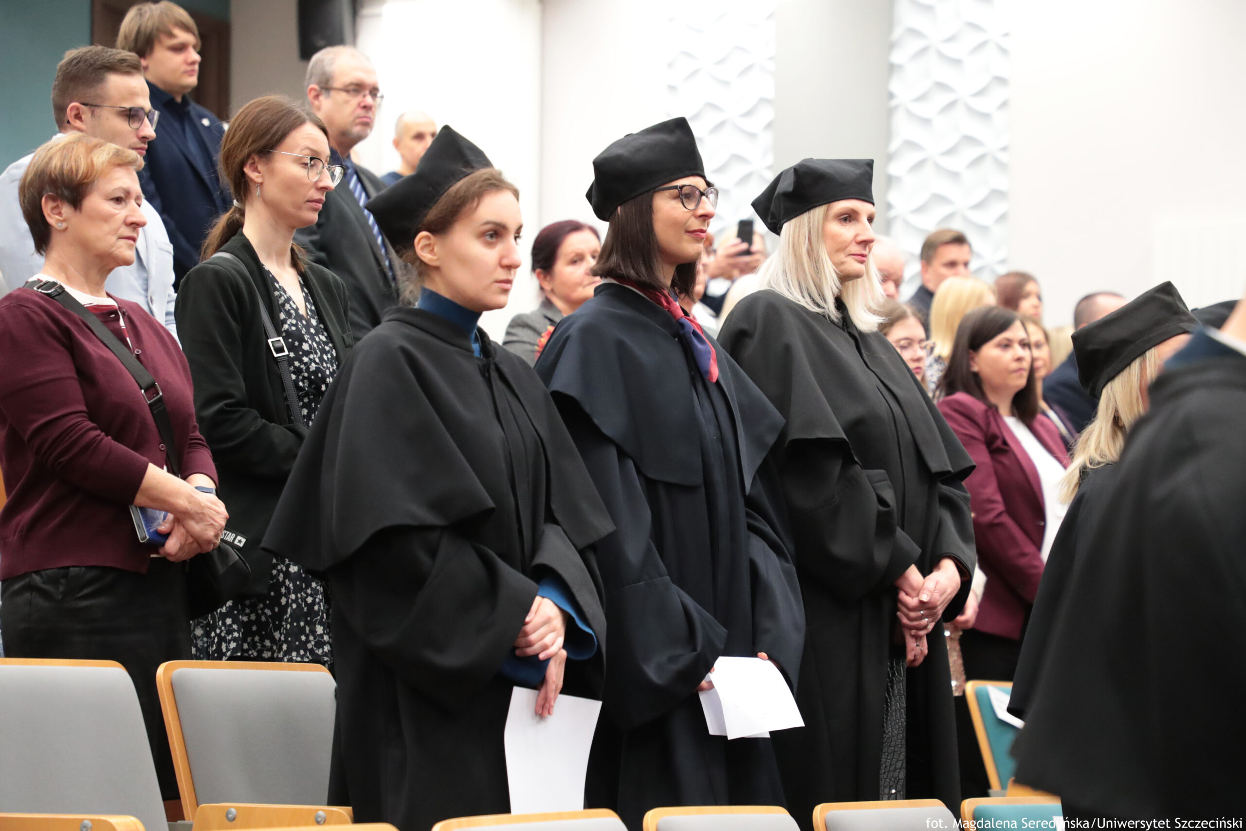 Grupa ludzi stojących w auli podczas uroczystości akademickiej. Na pierwszym planie stoją trzy kobiety w czarnych togach i czarnych biretach; dwie mają okulary. Dwie z nich trzymają w dłoniach białe kartki. Obok nich stoją inni uczestnicy w ubraniach codziennych, a w tle widać więcej osób, część siedzi na krzesłach. Wnętrze jest jasne, ze ścianami z dekoracyjnymi, wypukłymi panelami. W prawym dolnym rogu jest mały napis ze stopką zdjęcia, ale jest zbyt mały, by go pewnie przepisać.