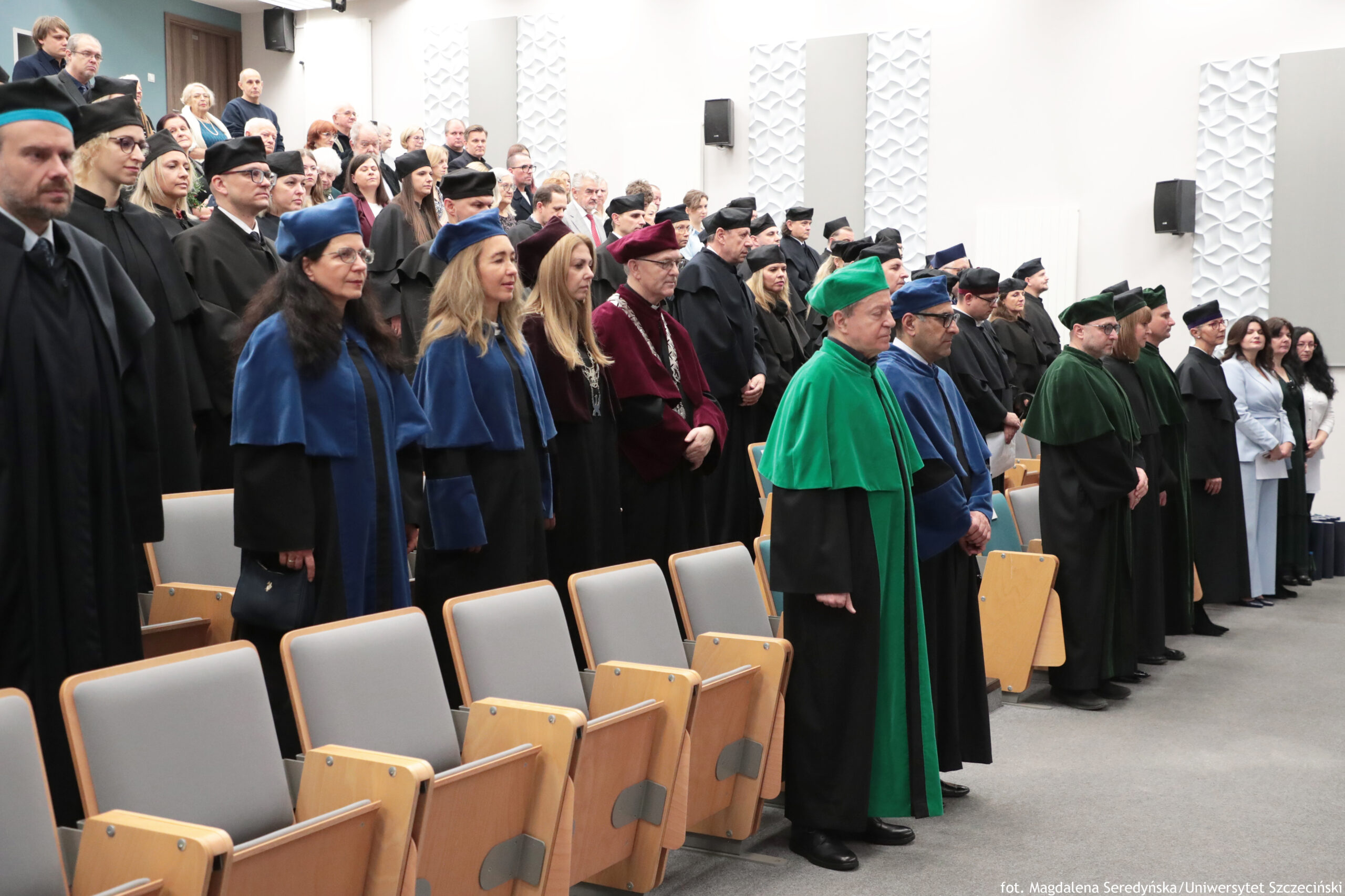 Grupa ludzi w auli uniwersyteckiej, stojąca w kilku rzędach, głównie w togach akademickich i biretach. W pierwszym planie widać rzędy jasnych, drewniano-tapicerowanych krzeseł. Wśród osób na pierwszym planie są togi w kolorach czarnym, granatowym, zielonym i bordowym; część osób ma łańcuchy dziekańskie. Większość osób to dorośli w średnim lub starszym wieku, o jasnej karnacji; stoją poważnie, zwróceni w jednym kierunku, jak podczas uroczystości. Sala ma białe ściany z dekoracyjnymi panelami i zamontowane głośniki. W prawym dolnym rogu jest mały napis: „fot. Magdalena Seredyńska/Uniwersytet Szczeciński”.