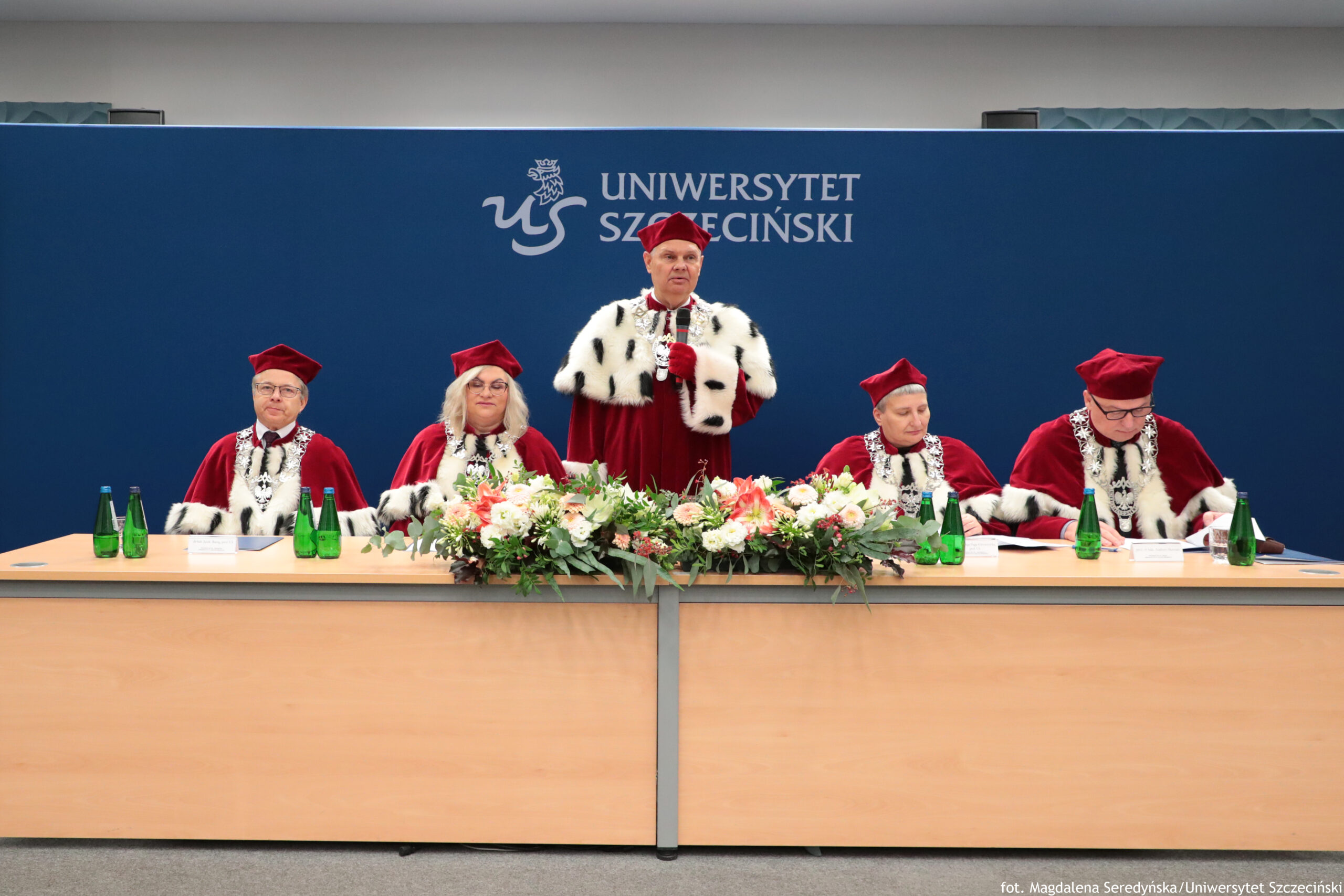 Grupa pięciu osób w czerwonych togach i czerwonych biretach przy długim stole, podczas uroczystości akademickiej. W tle jest granatowa ścianka z białym logo „US” i napisem „UNIWERSYTET SZCZECIŃSKI”. Osoba stojąca na środku (mężczyzna w średnim wieku, jasna skóra) przemawia, trzymając mikrofon. Cztery pozostałe osoby siedzą: po lewej mężczyzna w okularach, obok kobieta w okularach z jasnymi włosami; po prawej kobieta, a na skraju prawym mężczyzna w okularach, pochylony nad dokumentami. Stroje mają białe futrzane peleryny z czarnymi plamkami i ozdobne łańcuchy na piersi. Na stole stoi duży, podłużny bukiet kwiatów (białe, różowe i czerwone) z zielenią. Przy miejscach leżą kartki i dokumenty, stoją też zielone butelki wody. W prawym dolnym rogu widoczny jest podpis: „fot. Magdalena Seredyńska/Uniwersytet Szczeciński”.