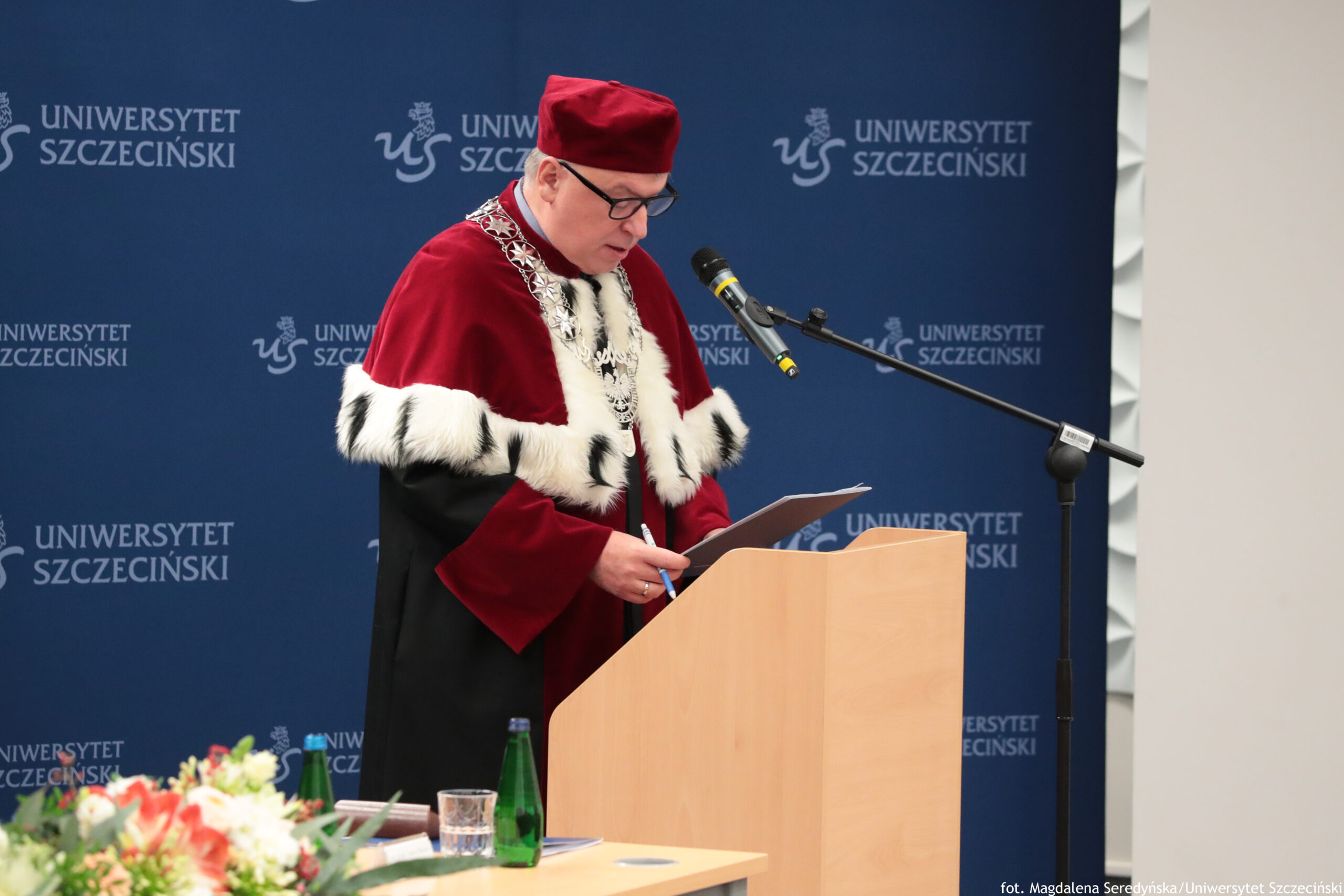 Na zdjęciu widać mężczyznę o jasnej karnacji w okularach, w uroczystym stroju akademickim: bordowa czapka i bordowo‑czarna toga z białym futrzanym wykończeniem oraz ozdobny łańcuch na szyi. Stoi przy jasnej drewnianej mównicy i czyta z kartki do mikrofonu na statywie. W tle jest granatowa ścianka z powtarzającym się napisem „UNIWERSYTET SZCZECIŃSKI” i logo. Na dole z przodu widać rozmyty bukiet kwiatów, zieloną butelkę i szklankę. W prawym dolnym rogu jest podpis: „fot. Magdalena Seredyńska/Uniwersytet Szczeciński”.
