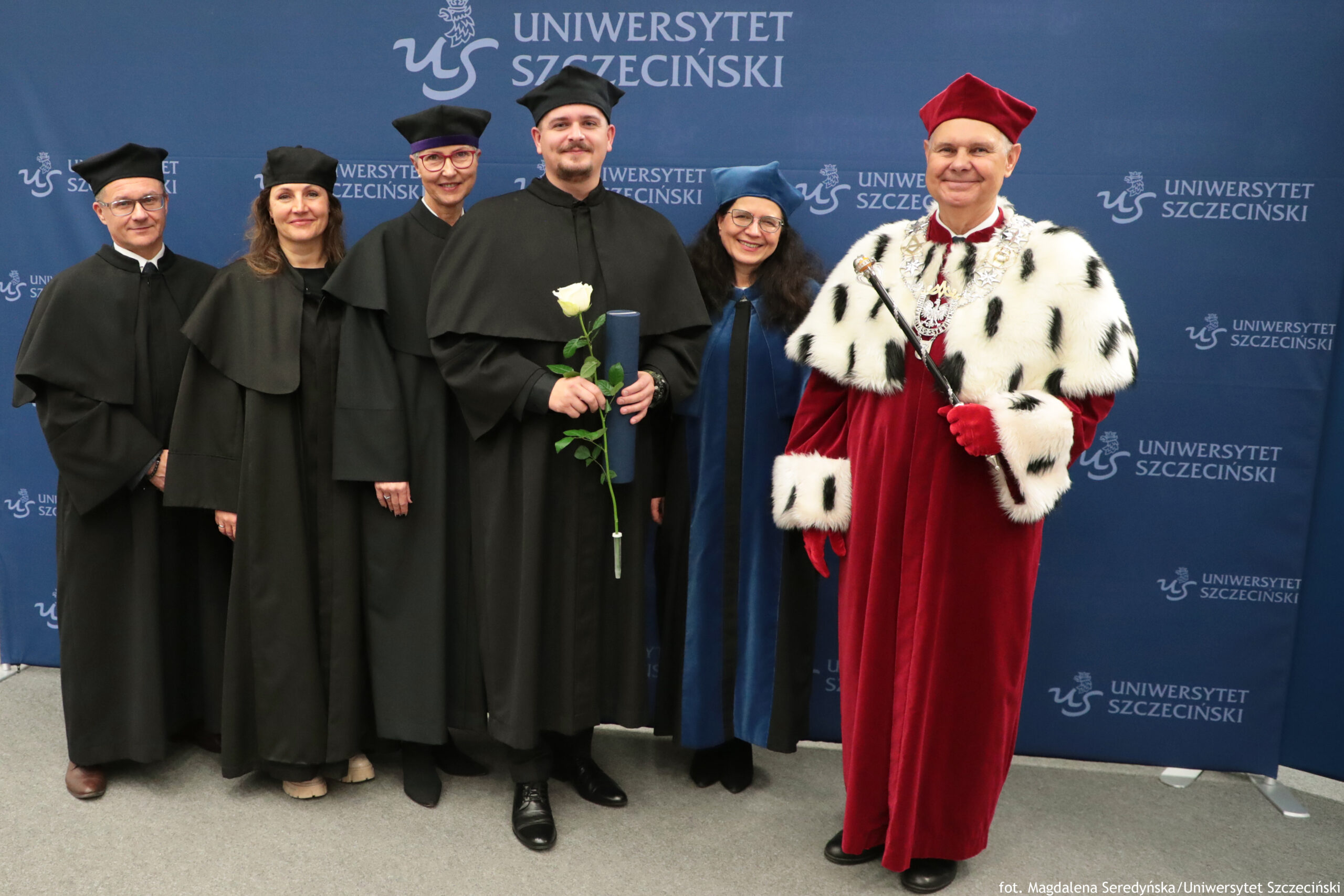 Grupowe zdjęcie sześciu dorosłych osób w strojach akademickich na tle ścianki z napisem „UNIWERSYTET SZCZECIŃSKI” i logo uczelni. Pięć osób stoi po lewej stronie w czarnych togach i biretach; jedna z nich ma czapkę i elementy stroju w kolorze niebieskim. Osoba stojąca w środku trzyma białą różę oraz granatową tubę na dyplom. Po prawej stronie stoi mężczyzna w ozdobnym, długim, czerwonym stroju z białym futrzanym płaszczem w czarne plamki, w czerwonej czapce i czerwonych rękawiczkach; trzyma berło. Na dole po prawej widać mały napis: „fot. Magdalena Seredyńska/Uniwersytet Szczeciński”.