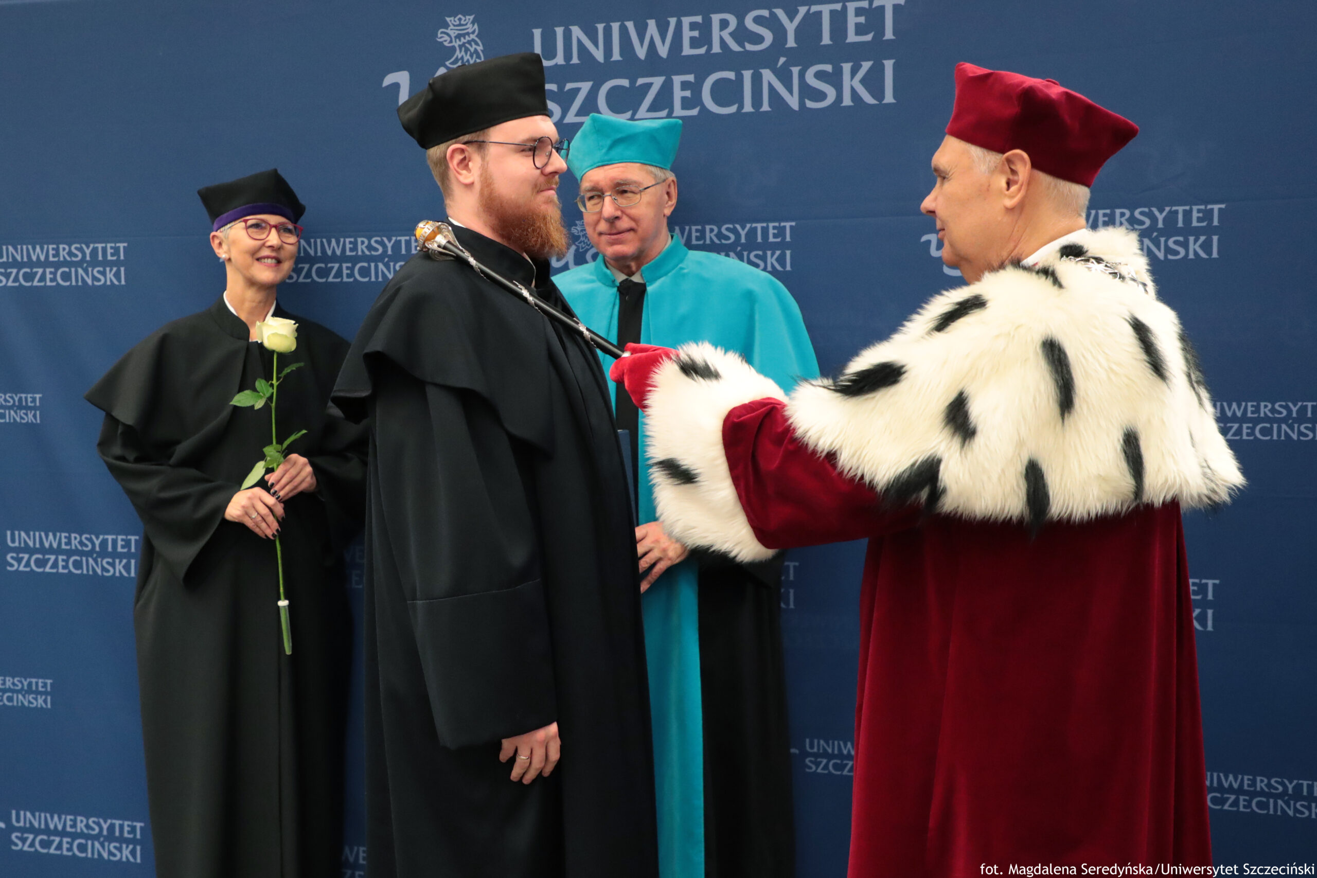 Ceremonia akademicka z czterema osobami w togach na tle ścianki z napisem „UNIWERSYTET SZCZECIŃSKI”. W centrum stoi mężczyzna o jasnej skórze z rudą brodą, w czarnej todze i czarnym birecie, w okularach. Z prawej strony stoi starszy mężczyzna o jasnej skórze w bordowej czapce i bordowej todze z białym futrzanym kołnierzem w czarne plamy; trzyma w ręku ozdobne insygnium i dotyka nim ramienia mężczyzny w czerni, jak podczas aktu promocji lub pasowania. Za nimi stoi mężczyzna o jasnej skórze w turkusowej todze i turkusowej czapce, również w okularach. Po lewej stoi kobieta o jasnej skórze w czarnej todze i czarnej czapce z fioletowym paskiem, w okularach; trzyma białą różę. Widoczny tekst w tle: „UNIWERSYTET SZCZECIŃSKI”. W prawym dolnym rogu: „fot. Magdalena Seredyńska/Uniwersytet Szczeciński”.